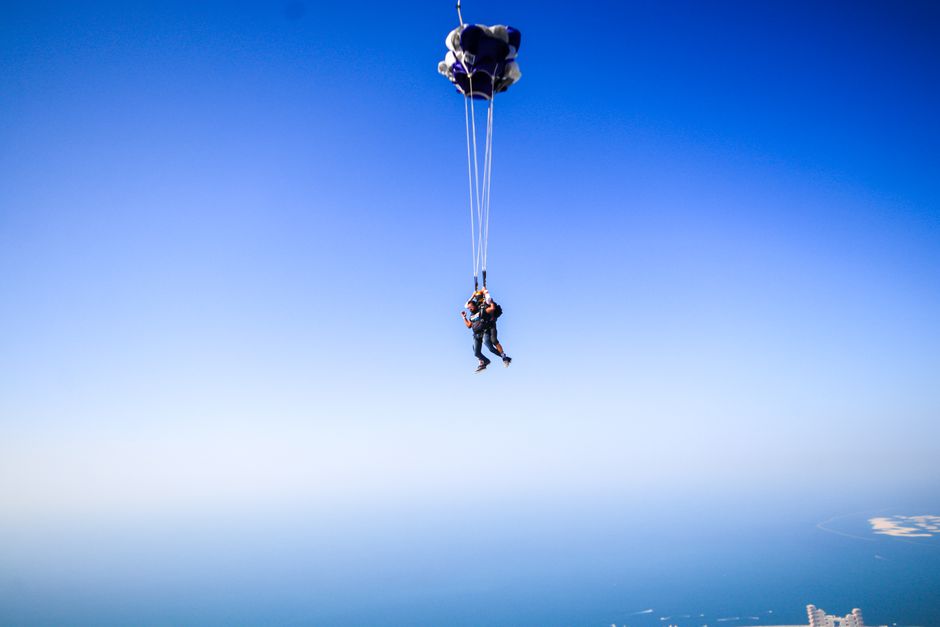 Two skydivers floating with parachutes over the calm turquoise ocean under a clear blue sky.