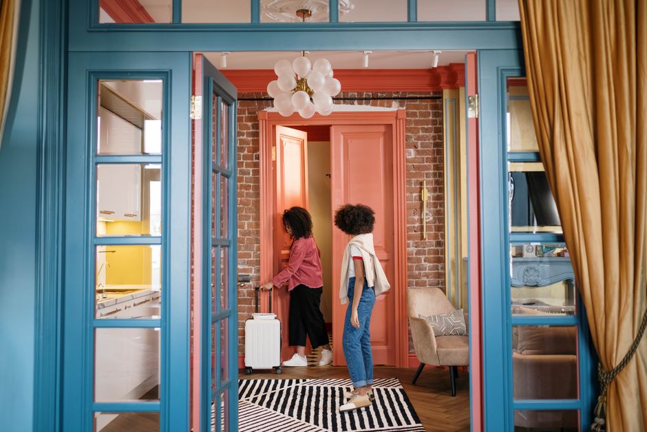 Stylish room with two women walking through doorway, carrying suitcase. Cozy and colorful decor.