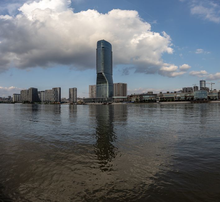 Urban skyline with a modern skyscraper reflecting in the river under a cloudy sky.