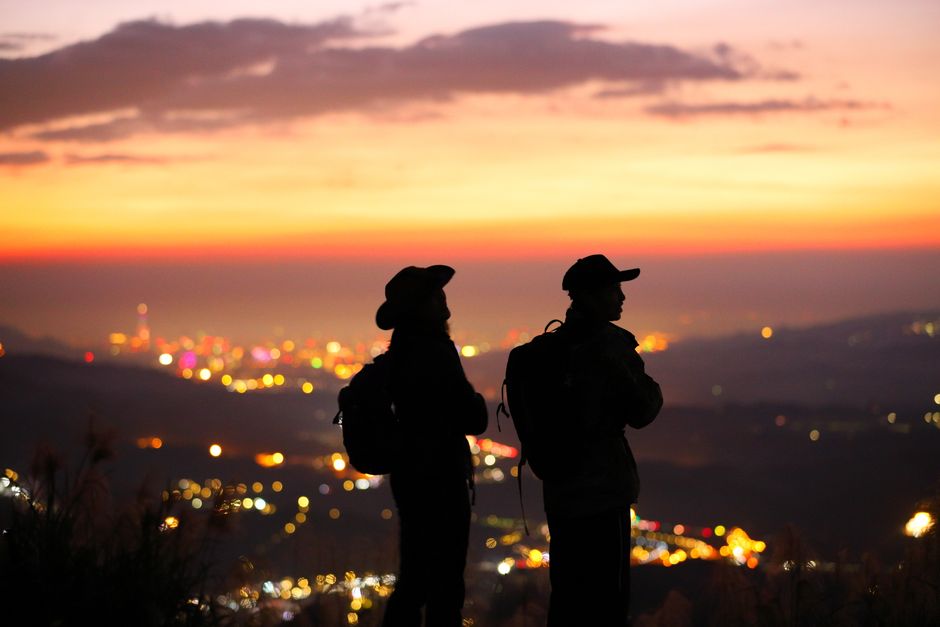 Two hikers silhouetted against a vibrant sunset over Taipei, creating a serene and adventurous scene.
