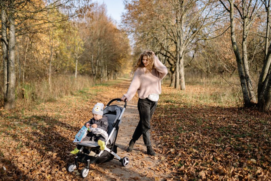 A mother walks with her child in a stroller along a leaf-covered path in a serene autumn park.