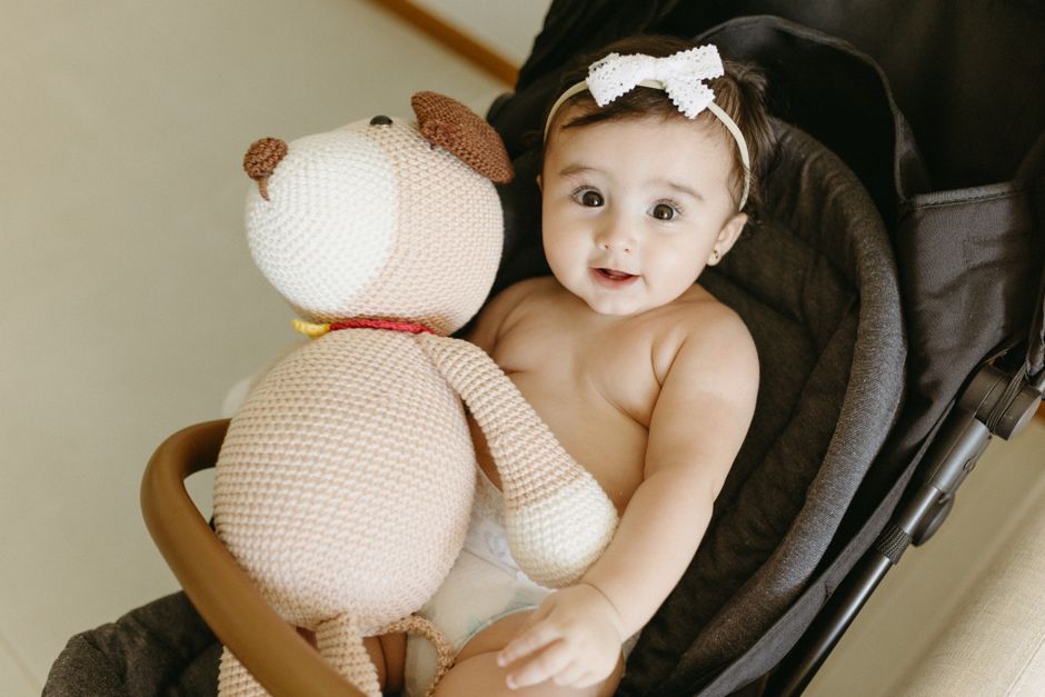 A cute baby in a stroller holding a knitted teddy bear inside a cozy indoor setting.