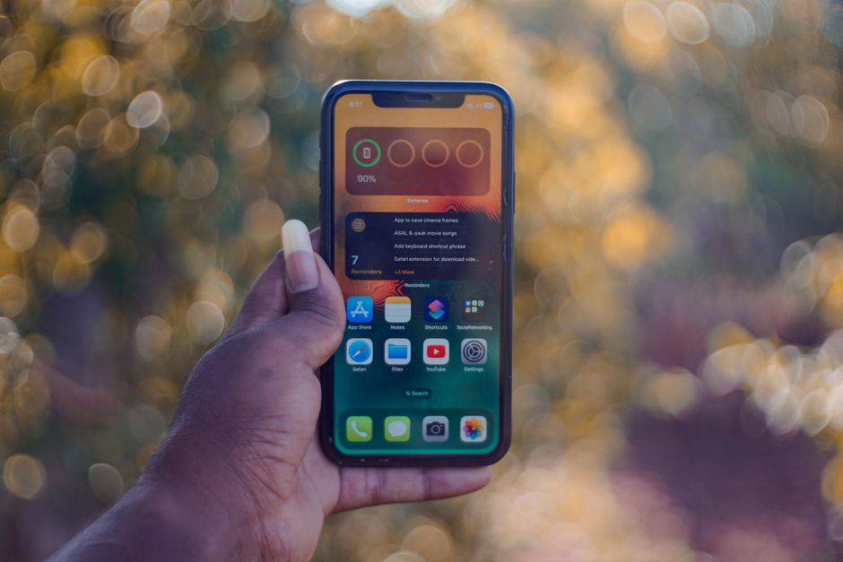 A hand holding a smartphone showing home screen apps, outdoors with bokeh background.