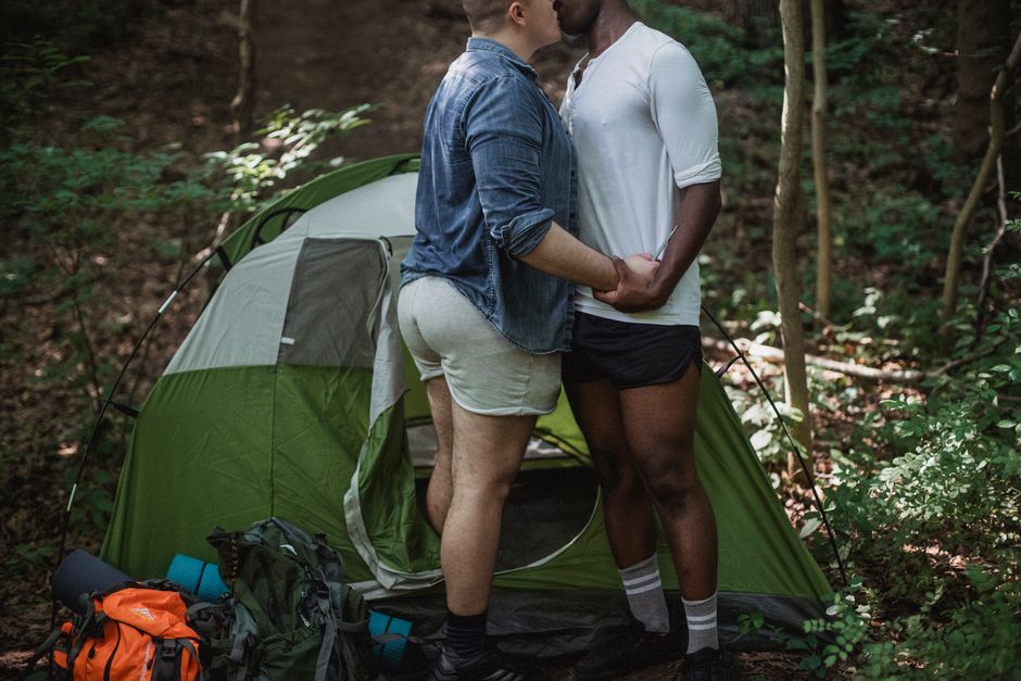 Crop of unrecognizable multiracial gay couple kissing on nature during summer vacation in forest in green tent