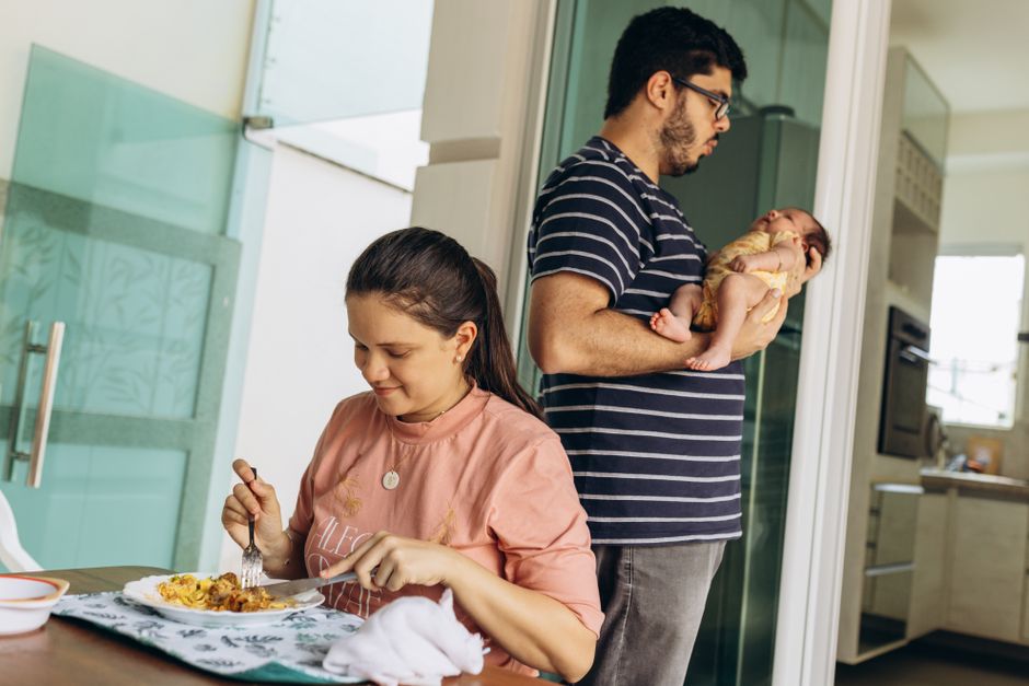 A woman enjoys a meal while a man cradles a baby, capturing family life.