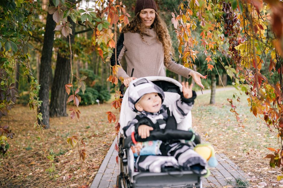 Mother walks with toddler in stroller through a scenic autumn park, surrounded by vibrant fall foliage.