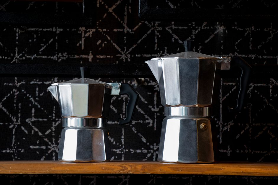 Close-up of two silver Italian stovetop espresso makers on a wooden shelf with a black patterned background.