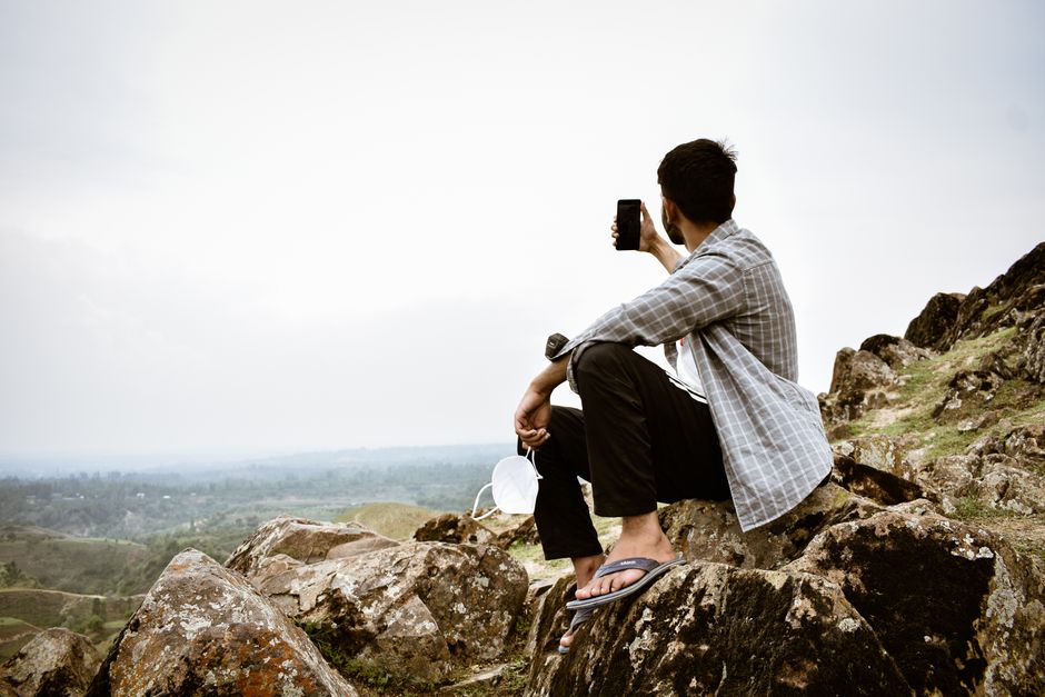 Man sits on rocky terrain capturing a selfie with scenic mountain background.
