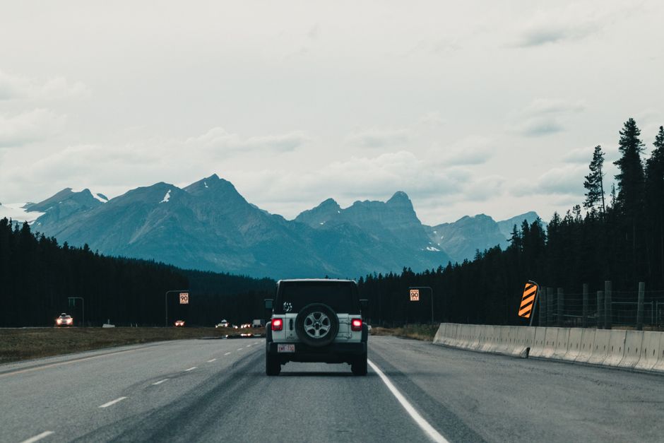 An SUV drives through a mountain highway with scenic views of towering peaks and forests.