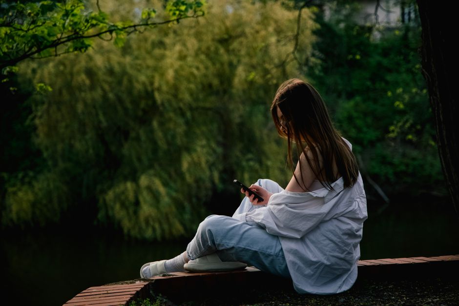 A young woman sits on a brick ledge in a lush green park, browsing her smartphone, creating a peaceful outdoor scene.