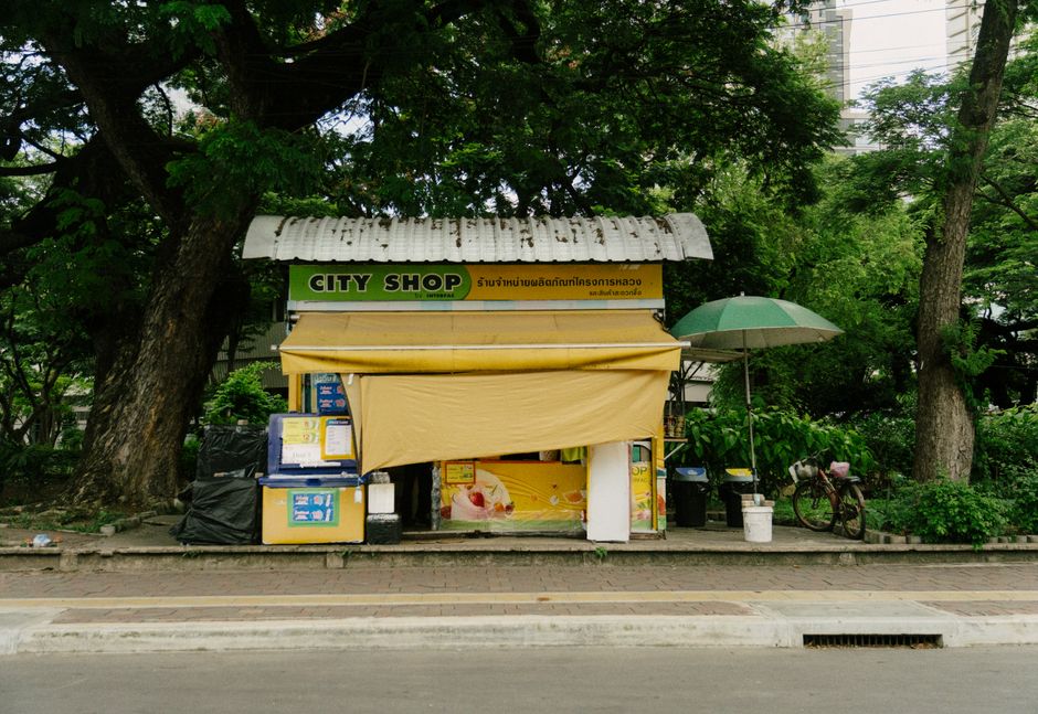 Charming street shop in Bangkok with lush greenery, featuring a yellow awning and colorful signage.